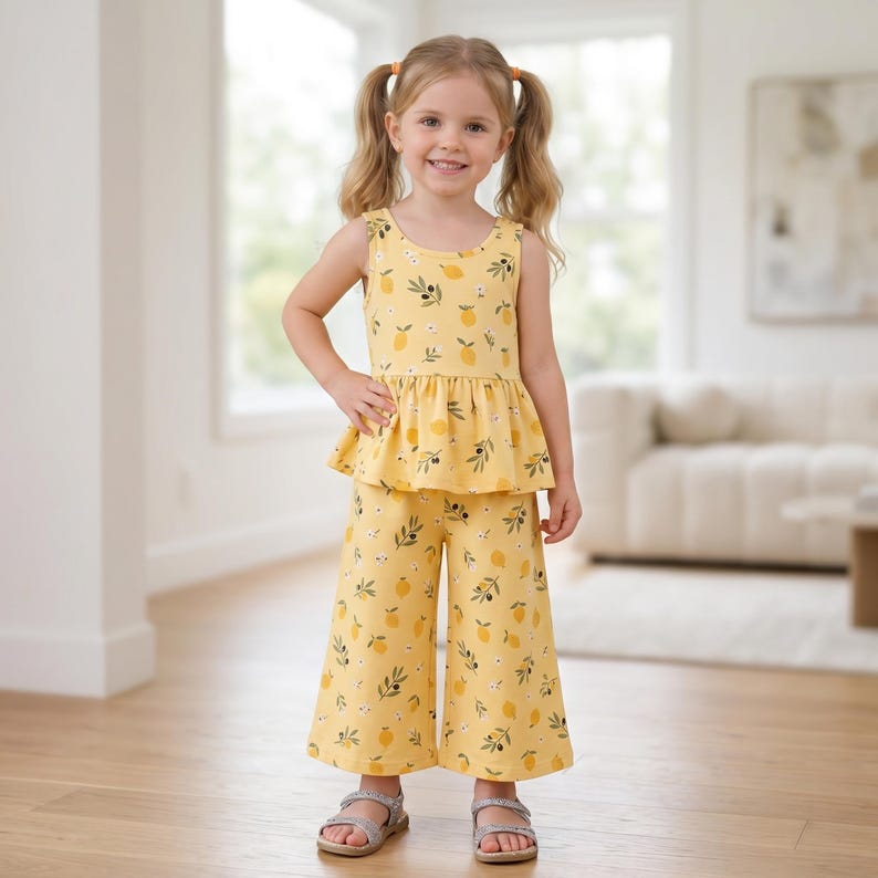 Op de afbeelding: Een tweedelige outfit bestaande uit een mouwloze peplumtop en een wijde broek, beide met een gele achtergrond en een herhalend patroon van kleine citroenen, groene blaadjes en kleine witte bloemetjes, afgewerkt met een ronde halslijn.