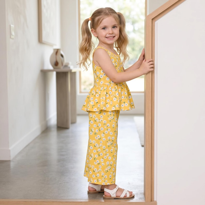 Op de afbeelding: Een tweedelige outfit bestaande uit een mouwloze peplumtop en een bijpassende broek met wijde pijpen, beide met een felgele achtergrond en een herhalend patroon van kleine witte madeliefjes met gele hartjes, gecombineerd met witte sandalen.