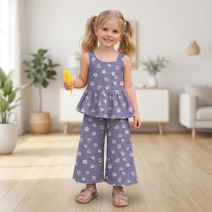 Op de afbeelding: Een tweedelige set bestaande uit een mouwloze peplumtop en een wijde broek, beide voorzien van een herhalend bloemenpatroon van witte en roze rozen op een gedempte paarse achtergrond. De top heeft een ronde halslijn en een uitlopende taille.
