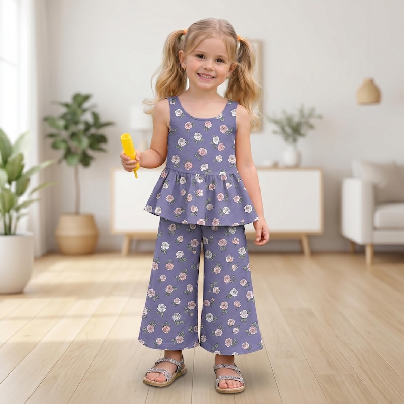 Op de afbeelding: Een tweedelige set bestaande uit een mouwloze peplumtop en een wijde broek, beide voorzien van een herhalend bloemenpatroon van witte en roze rozen op een gedempte paarse achtergrond. De top heeft een ronde halslijn en een uitlopende taille.