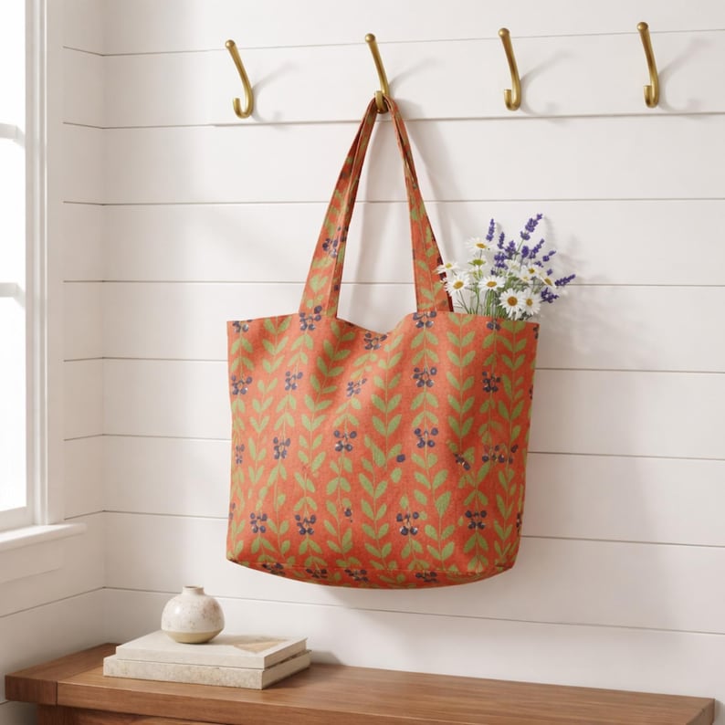 May include: An orange tote bag with a green and blue leaf pattern hangs from a brass hook. The bag is filled with white daisies and purple lavender. The bag is hanging on a white wall with horizontal wood paneling.