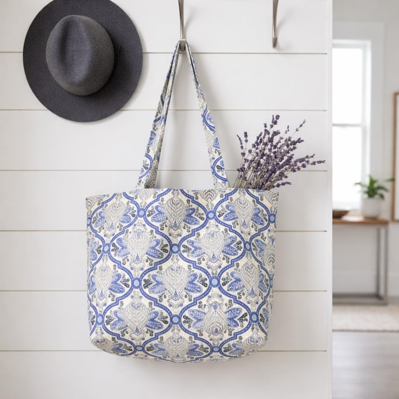 May include: A tote bag with a blue and white floral pattern hangs on a wall. The bag is filled with lavender. A gray fedora hat hangs on the wall to the left of the bag.