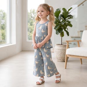Op de afbeelding: Een set bestaande uit een mouwloze peplumtop en een bijpassende wijde, kortere broek in stofblauw met een herhalend patroon van witte meeuwen en kleine zwarte ankers. De outfit wordt gecombineerd met eenvoudige witte sandalen met bandjes.