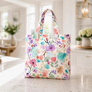 May include: A floral tote bag with a white background and colorful watercolor flowers in shades of pink, purple, and teal. The bag has matching handles and is sitting on a marble countertop.