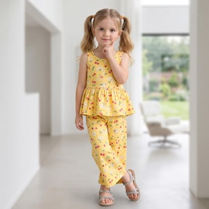 Op de afbeelding: Een gele mouwloze peplumtop en bijpassende broek met wijde pijpen, voorzien van een herhalend patroon van kleine rode kersen en witte madeliefjes. De outfit wordt gedragen met zilverkleurige sandalen met bandjes en een gespsluiting.