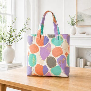 May include: A colorful tote bag with a cream background and abstract shapes in orange, purple, teal, and gray. The bag has matching handles and gold accents. The tote bag is sitting on a wooden table.
