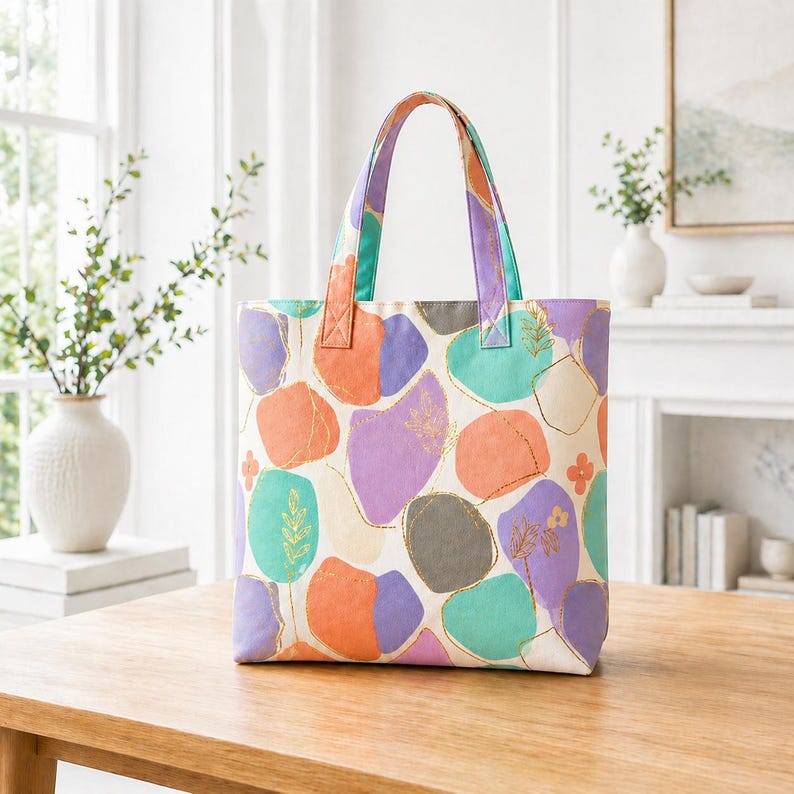 May include: A colorful tote bag with a cream background and abstract shapes in orange, purple, teal, and gray. The bag has matching handles and gold accents. The tote bag is sitting on a wooden table.