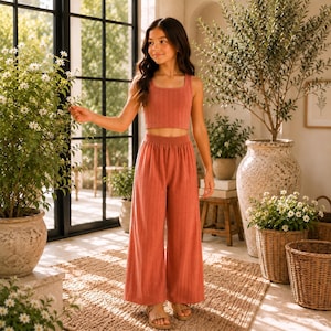 May include: A two-piece set consisting of a sleeveless, ribbed crop top and matching wide-leg trousers in a solid terracotta colour. The garment features a vertical textured pattern throughout, with a simple scoop neckline and a high-waisted fit.