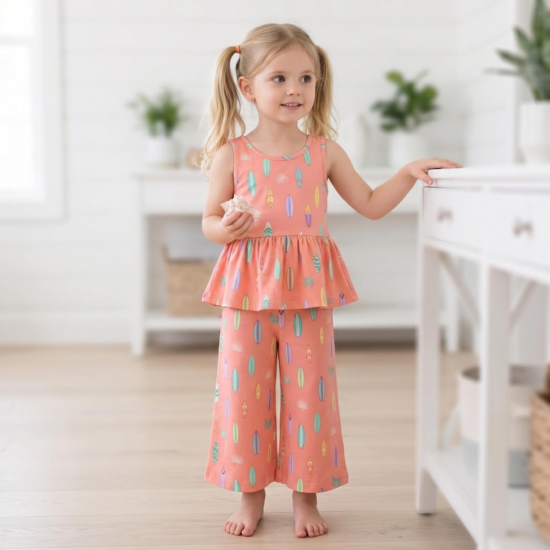 Op de afbeelding: Een perzikkleurige mouwloze peplumtop en bijpassende broek met wijde pijpen, voorzien van een all-over print van kleurrijke surfplanken in tinten turquoise, geel en paars. De set is gemaakt van een lichtgewicht stof met een ronde halslijn.