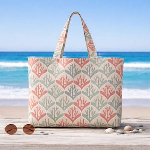 May include: A beige tote bag with a repeating coral reef pattern in shades of pink and green. The bag has matching handles and is set against a beach backdrop with sunglasses and pebbles.