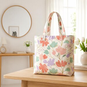 May include: A floral tote bag with a cream background and a pattern of pink, coral, lavender, and teal flowers. The bag has matching handles and is sitting on a wooden table.