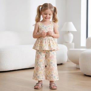 Op de afbeelding: Een tweedelige outfit bestaande uit een mouwloze peplumtop en een wijde broek, beide met een cr&egrave;mekleurige basis en een herhalend bloemenpatroon in zachte tinten roze, blauw en geel, gecombineerd met eenvoudige grijze sandalen met bandjes.