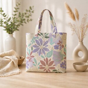 May include: A floral tote bag with a cream background and a pattern of purple, pink, and green flowers and leaves. The bag has two handles and is sitting on a wooden surface. The bag is a square shape.