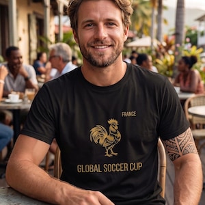Pode incluir: Camiseta preta com um galo dourado e o texto "FRANCE" e "GLOBAL SOCCER CUP". A pessoa que usa a camiseta está sentada em uma mesa ao ar livre. A pessoa tem uma tatuagem no braço.
