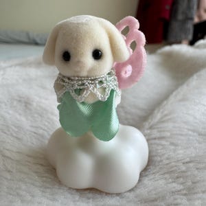 Puede incluir: Una peque&ntilde;a figura de perro color crema con ojos negros, que lleva un collar de encaje plateado y una cinta verde menta. Tiene alas de fieltro rosa y se asienta sobre una base blanca en forma de nube. Un art&iacute;culo decorativo caprichoso.