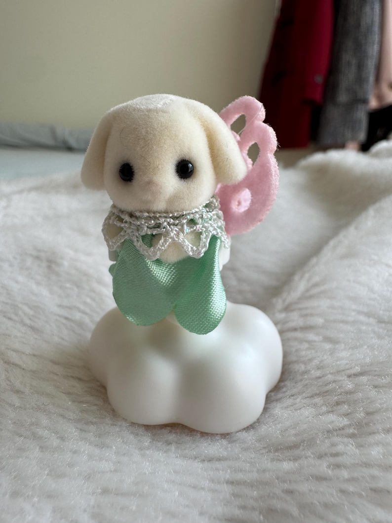 Puede incluir: Una peque&ntilde;a figura de perro color crema con ojos negros, que lleva un collar de encaje plateado y una cinta verde menta. Tiene alas de fieltro rosa y se asienta sobre una base blanca en forma de nube. Un art&iacute;culo decorativo caprichoso.