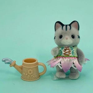 Puede incluir: Una figura de gato gris con un vestido verde y rosa con una falda en forma de flor. Junto al gato hay una regadera beige con un dise&ntilde;o floral y una boquilla plateada. El fondo es azul claro.