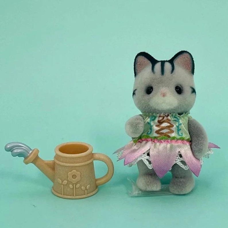 Puede incluir: Una figura de gato gris con un vestido verde y rosa con una falda en forma de flor. Junto al gato hay una regadera beige con un dise&ntilde;o floral y una boquilla plateada. El fondo es azul claro.