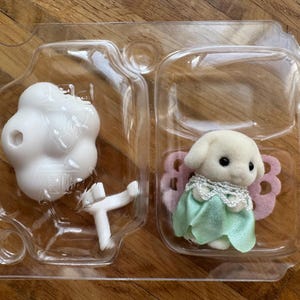 Puede incluir: Un perro de juguete blanco con un vestido verde y alas rosas, empaquetado en un recipiente de pl&aacute;stico transparente. El conjunto incluye un objeto blanco en forma de nube y un soporte blanco. El juguete es para ni&ntilde;os.