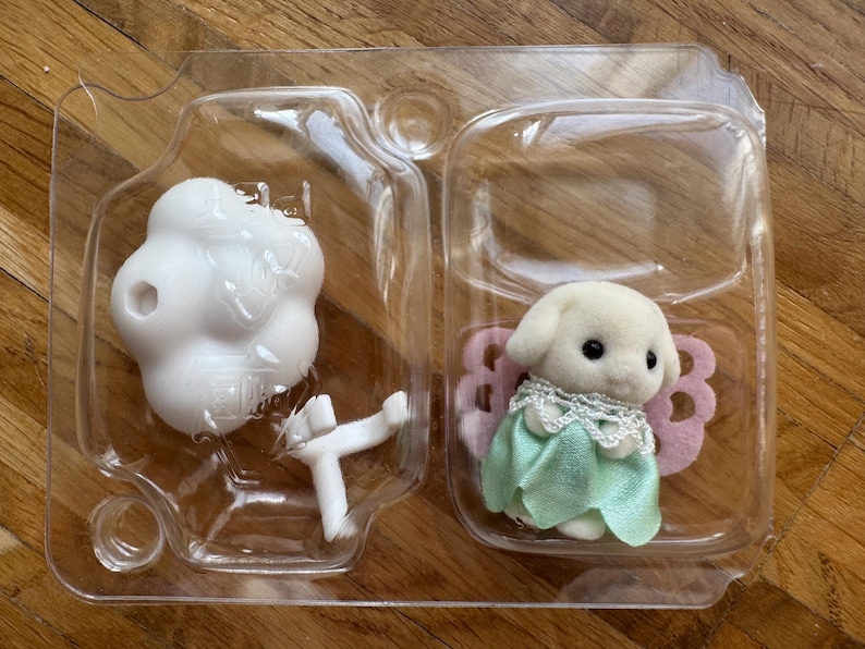 Puede incluir: Un perro de juguete blanco con un vestido verde y alas rosas, empaquetado en un recipiente de pl&aacute;stico transparente. El conjunto incluye un objeto blanco en forma de nube y un soporte blanco. El juguete es para ni&ntilde;os.