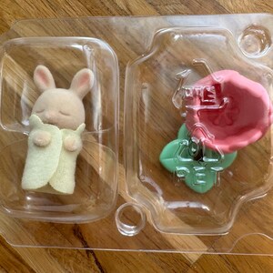 Puede incluir: Una peque&ntilde;a figura de conejito beige con los ojos cerrados, envuelta en una manta verde claro, y un accesorio en forma de flor rosa, empaquetada en un recipiente de pl&aacute;stico transparente. El conejito tiene orejas rosas.
