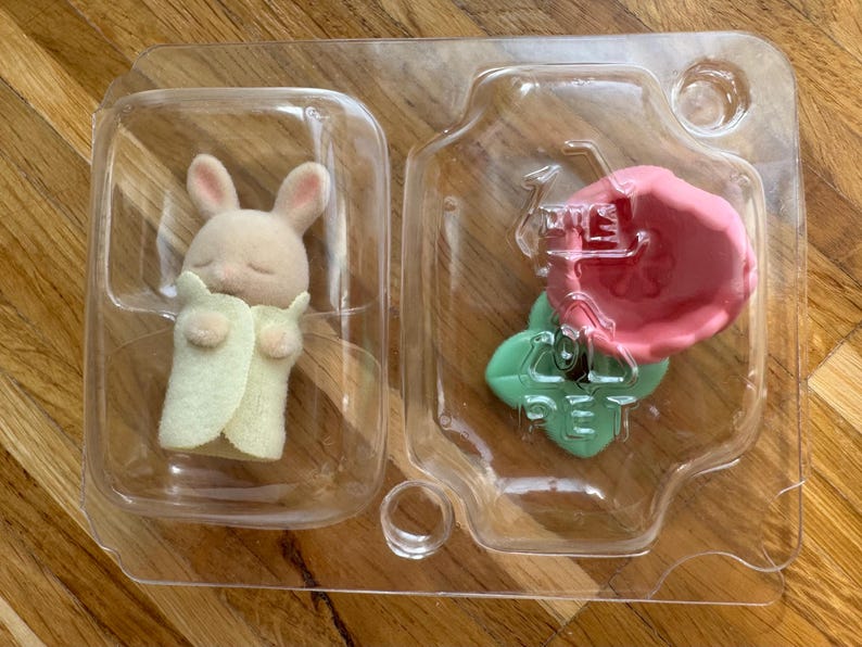 Puede incluir: Una peque&ntilde;a figura de conejito beige con los ojos cerrados, envuelta en una manta verde claro, y un accesorio en forma de flor rosa, empaquetada en un recipiente de pl&aacute;stico transparente. El conejito tiene orejas rosas.