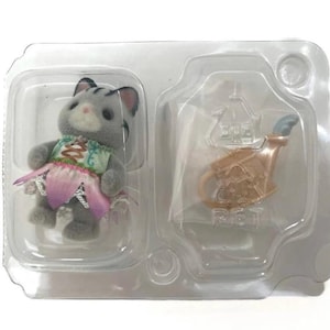 Puede incluir: Una figura de gato gris con un vestido colorido con una falda rosa y una peque&ntilde;a regadera en un paquete de pl&aacute;stico transparente. El gato tiene rayas negras y una nariz rosa.