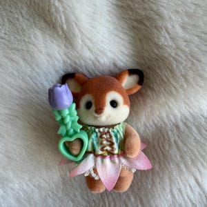 Puede incluir: Peque&ntilde;a figura de zorro marr&oacute;n y blanco con un vestido verde y rosa con una falda en forma de flor. El zorro sostiene una flor morada con un tallo verde y un asa en forma de coraz&oacute;n. La figura est&aacute; sobre una superficie blanca y esponjosa.