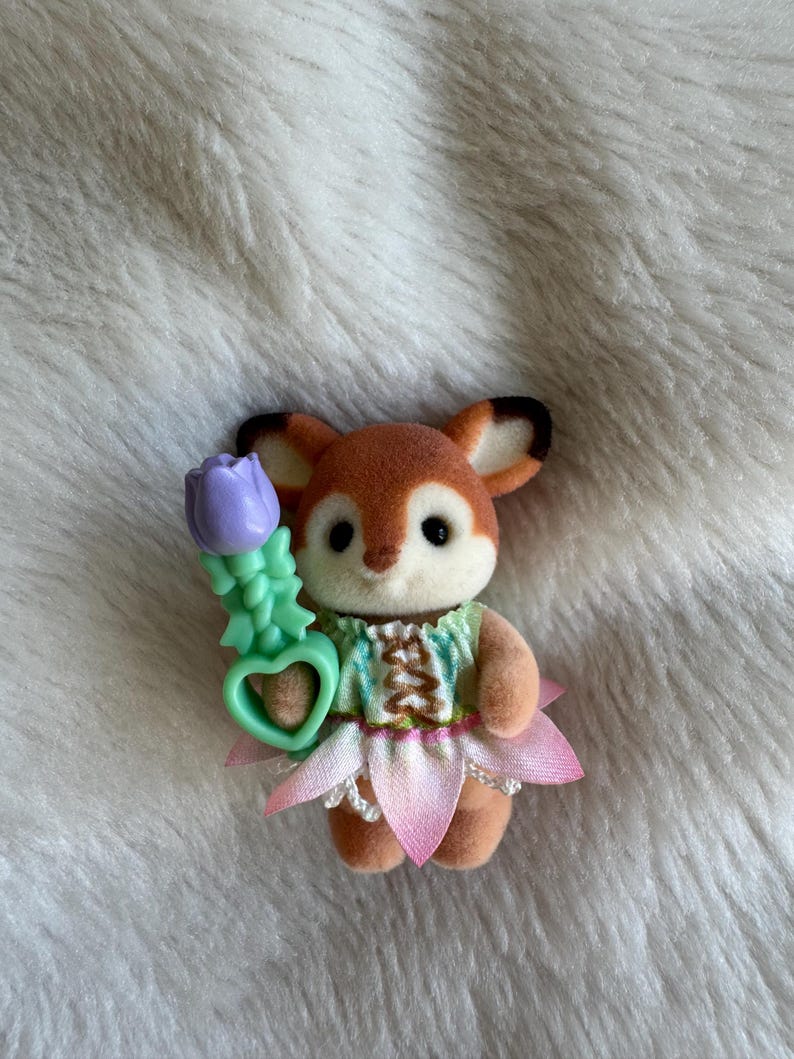 Puede incluir: Peque&ntilde;a figura de zorro marr&oacute;n y blanco con un vestido verde y rosa con una falda en forma de flor. El zorro sostiene una flor morada con un tallo verde y un asa en forma de coraz&oacute;n. La figura est&aacute; sobre una superficie blanca y esponjosa.