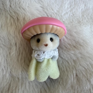Puede incluir: Una peque&ntilde;a figura en forma de hongo gris con una tapa rosa y una parte inferior beige. Tiene ojos negros y una peque&ntilde;a nariz marr&oacute;n. La figura lleva un cuello de encaje blanco y una capa verde claro.