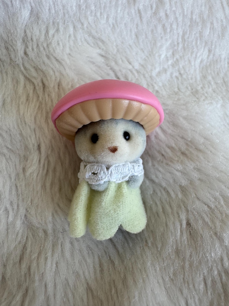Puede incluir: Una peque&ntilde;a figura en forma de hongo gris con una tapa rosa y una parte inferior beige. Tiene ojos negros y una peque&ntilde;a nariz marr&oacute;n. La figura lleva un cuello de encaje blanco y una capa verde claro.