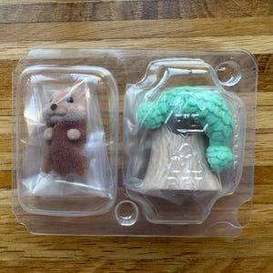Puede incluir: Un paquete de pl&aacute;stico transparente que contiene dos peque&ntilde;as figuras. Una es una criatura marr&oacute;n y beige parecida a una ardilla. La otra es un &aacute;rbol con una parte superior verde texturizada. Las figuras son probablemente juguetes o art&iacute;culos de colecci&oacute;n.