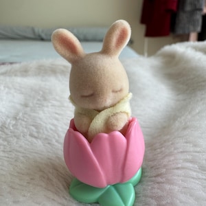 Puede incluir: Una peque&ntilde;a figura de conejito beige dentro de un recipiente en forma de tulip&aacute;n rosa con hojas verdes. El conejito tiene los ojos cerrados y est&aacute; envuelto en una manta amarilla. Juguete infantil.