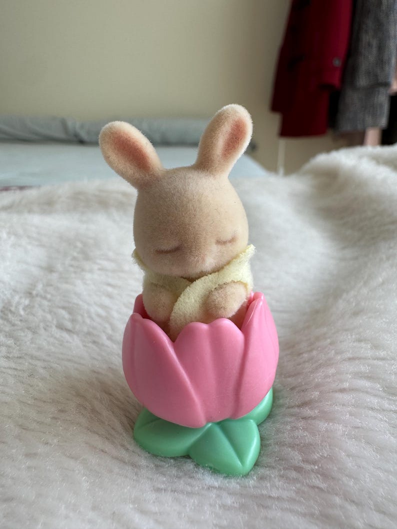 Puede incluir: Una peque&ntilde;a figura de conejito beige dentro de un recipiente en forma de tulip&aacute;n rosa con hojas verdes. El conejito tiene los ojos cerrados y est&aacute; envuelto en una manta amarilla. Juguete infantil.