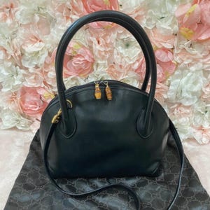 May include: A black leather handbag with a rounded silhouette, featuring two sturdy top handles and a detachable shoulder strap. The bag includes a top zipper closure with two distinct bamboo-style metal pulls and gold-toned hardware accents.