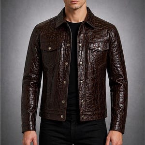May include: A brown leather jacket with a crocodile skin texture. The jacket has a button-up front, two chest pockets, and a collared neckline. The model is wearing a black shirt and black jeans. The jacket is a stylish outerwear option.
