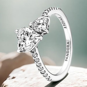 Può includere: Un anello d'argento con due pietre preziose chiare a forma di cuore. La fascia è impreziosita da piccole pietre chiare su un lato. L'anello è marchiato "S925 ALE" e "PANDORA".