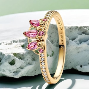 Puede incluir: Un anillo dorado con diseño de corona. La corona presenta tres piedras rosas en forma de marquesa y piedras rosas más pequeñas. La banda del anillo está adornada con pequeñas piedras preciosas transparentes. El texto "PANDORA ALE MET" está grabado en la banda.