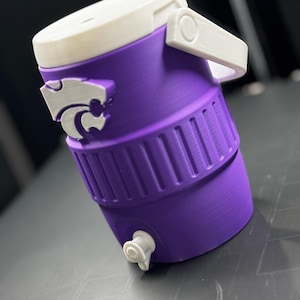 May include: A purple water cooler with a white lid and handle. The cooler features a silver logo and a white spigot. The design includes a ribbed band around the middle. This item is likely a sports or outdoor accessory.