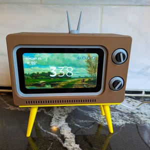 May include: A retro-style digital clock shaped like a vintage television. The brown clock has yellow legs and a screen displaying a landscape painting and the time. It features two knobs and an antenna.