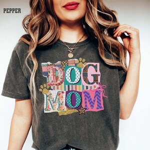Peut inclure: T-shirt gris foncé avec l'inscription "DOG MOM" dans un design patchwork coloré. Les lettres présentent divers motifs et couleurs, dont des motifs floraux, léopard et rayés. Des empreintes de pattes dorées à paillettes et une étoile ornent le design.