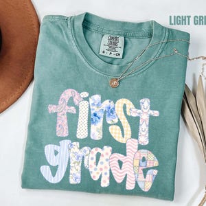 Peut inclure: Un t-shirt à manches courtes vert clair avec un col rond. L'avant présente le texte First Grade en grandes lettres de style bloc, chacune remplie d'un motif de tissu pastel différent, incluant des imprimés floraux, cachemire et vichy.
