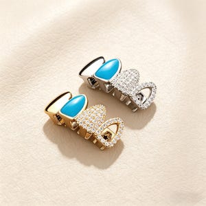 Op de afbeelding: Twee sets decoratieve tandgrills. De ene set is goudkleurig met een turquoise accent en een hartvormig ontwerp bezet met kleine, heldere kristallen. De andere set is zilverkleurig met een turquoise accent en een hartvormig ontwerp bezet met kleine, heldere kristallen.
