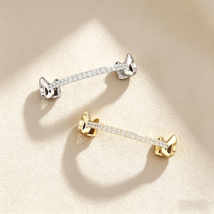 May include: Two decorative bar-shaped jewelry pieces. One is silver-toned, the other gold-toned. Each features a row of small, clear crystals set in a straight line. The ends have a stylized, curved design.
