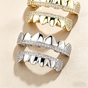May include: Close-up of three sets of gold and silver grillz. The top two sets are gold, encrusted with small, clear stones. The bottom two sets are silver, also with clear stones. The grillz are designed to fit over teeth.