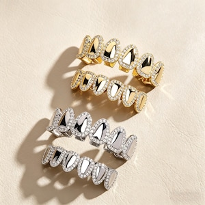 May include: Gold and silver-toned grillz with a diamond-like embellishment. The grillz have a row of pointed, oval-shaped teeth. The gold grillz are on top, and the silver grillz are on the bottom. The grillz are displayed on a neutral background.