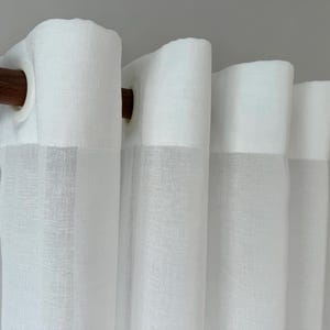 May include: A sheer white curtain panel with a subtle woven texture, featuring white grommets along the top header. The curtain is suspended from a dark brown wooden curtain rod, creating uniform vertical folds across the fabric surface.