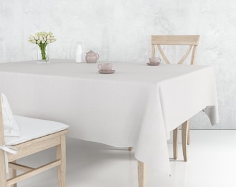 May include: A white linen tablecloth covers a wooden table, set with a vase of white flowers, a milk bottle, a teapot, and two teacups. Two wooden chairs are positioned around the table. The scene is set against a neutral, textured wall.
