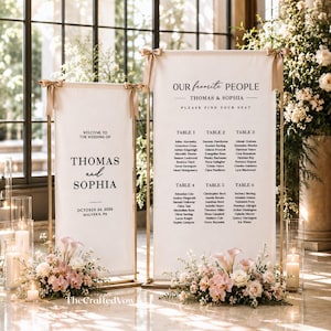 Peut inclure: Deux panneaux de mariage verticaux en tissu blanc avec des cadres en métal doré et des nœuds en ruban beige. Le panneau de gauche indique Welcome to the wedding of Thomas and Sophia, October 24, 2026, Malvern, PA. Le droit liste les tables.