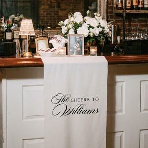May include: A wedding bar is decorated with a white table runner bearing the words "The Cheers to Williams". The bar is decorated with white flowers, framed photos, candles, and a small lamp. The background features a brick wall.
